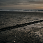 A single dark line across wet sand at dusk, with a distant horizon and faint amber sun.